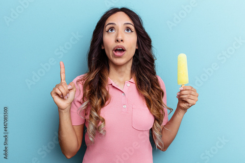 Young latin woman holding ice cream isolated on blue background pointing upside with opened mouth.