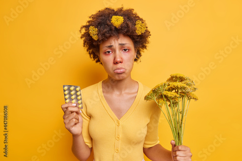 Photo of allergic curly haired woman looks sadly puses lips holds pills to treat allergy has unpleasant symptoms holds wildflowers picked from field isolated over yellow background. Monochrome photo