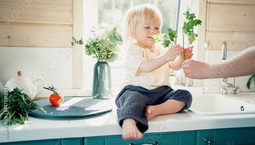 A beautiful child of European appearance, a blonde, plays with vegetables at home. A little boy is sitting on a table in the kitchen.