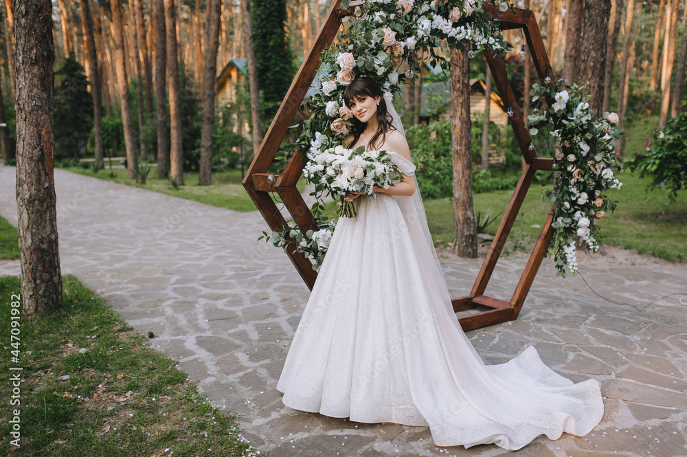 Fototapeta premium Wedding portrait of a beautiful, cute brunette bride in a white dress with a long veil with a large bouquet of flowers in nature in the park, forest, against the background of a decorated wooden arch.
