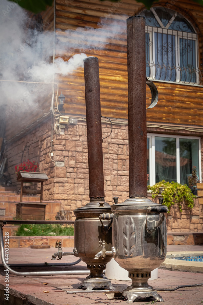 Two samovars with large pipes are fired with wood and release clouds of white smoke against the backdrop of a rural house Two samovars with large pipes are fired with wood and release clouds of white smoke against the backdrop of a rural house