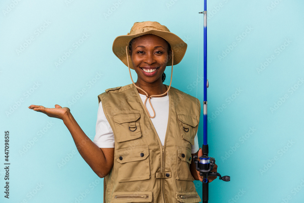 Young african american fisherwoman holding rod isolated on blue background showing a copy space on a palm and holding another hand on waist. Young african american fisherwoman holding rod isolated on blue background showing a copy space on a palm and holding another hand on waist.
