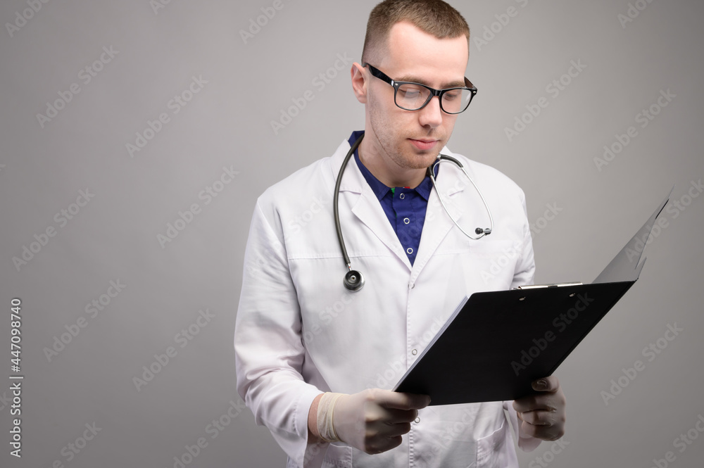 Portrait of a young male doctor in glasses with a stethoscope around his neck. In the hands of a folder, a tablet holder for papers. Studio shot on gray background Portrait of a young male doctor in glasses with a stethoscope around his neck. In the hands of a folder, a tablet holder for papers. Studio shot on gray background