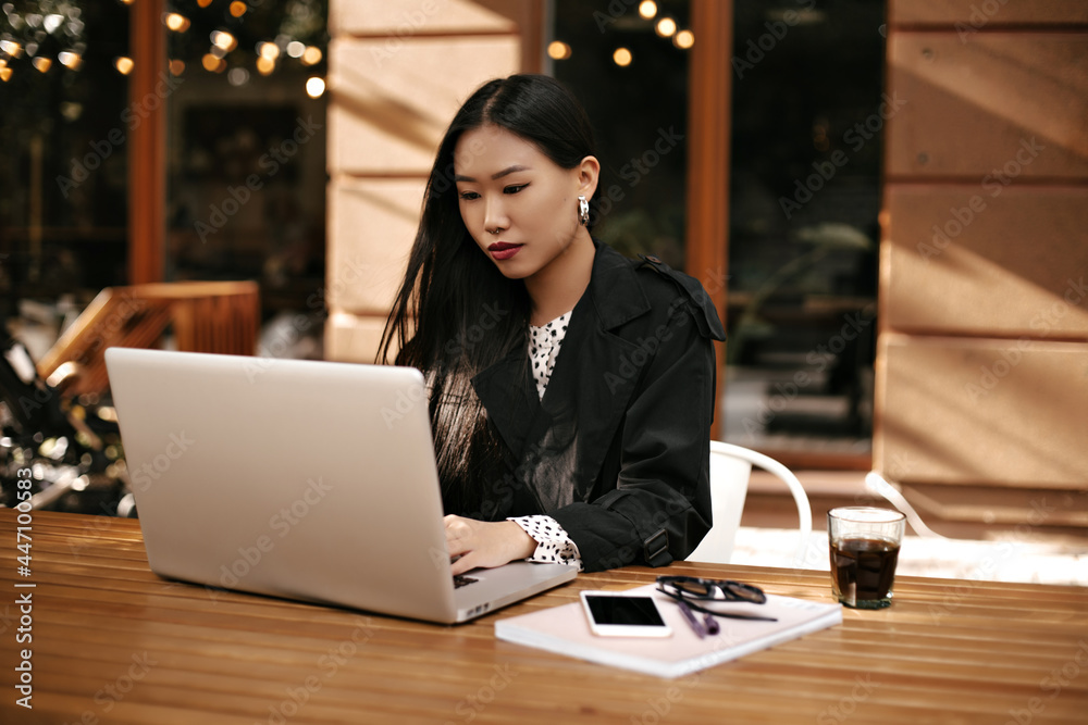 Brunette tanned woman in black jacket sits outside, works in computer. Smart lady in stylish outfit poses near wooden desk with coffee glass, phone and notebook. Brunette tanned woman in black jacket sits outside, works in computer. Smart lady in stylish outfit poses near wooden desk with coffee glass, phone and notebook.