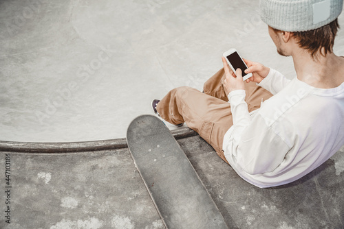 Teenager skater guy sits on the edge of a ramp in a gray skatepark near his skateboard and looks at the phone, checks messages, communicates with friends, browses social networks photo