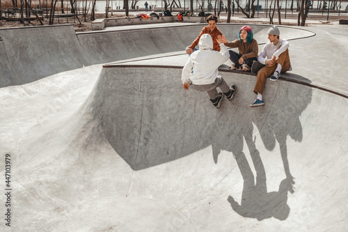 Group of teenagers hanging out in a city skatepark, rollerblading, sitting on a ramp and encouraging each other clapping their hands