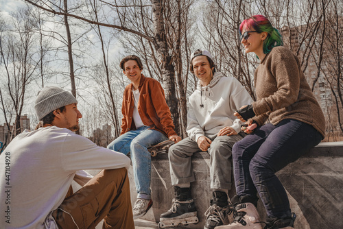 A group of subcultural teenagers, three boys and one girl, get-together in a spring / autumn urban skatepark in the city with rollerblades and skateboards, communicate with each other