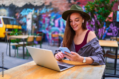 Happy cheerful smiling beautiful cute joyful young millennial freelancer girl wearing wireless headphones using laptop and phone for remote work, social networking and online browsing at cafe