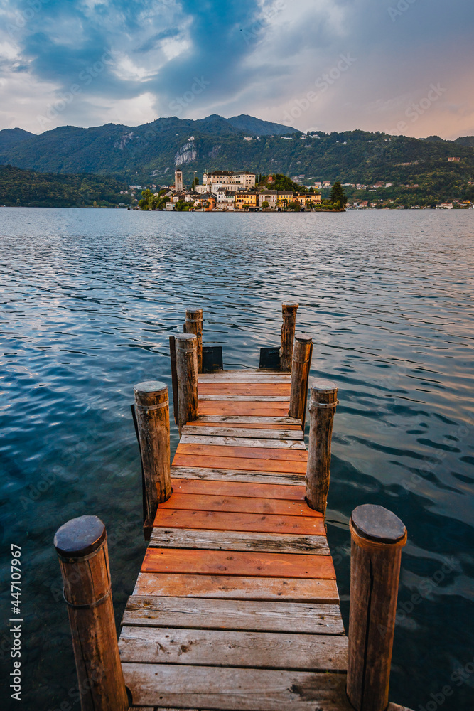 Naklejka premium Orta San Giulio / Italy - June 2021: The island of San Giulio with a wooden jetty in the foreground at sunset