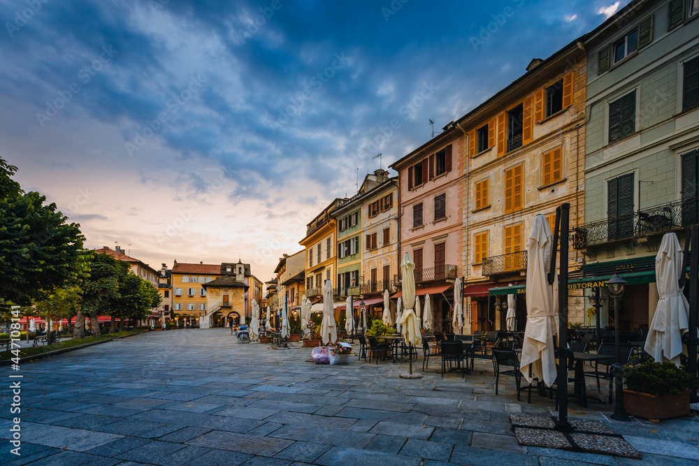 Orta San Giulio / Italy - June 2021: Main square of the village of Orta San Giulio at sunset with blue clouds, with people sitting in the bars / restaurants of the square
