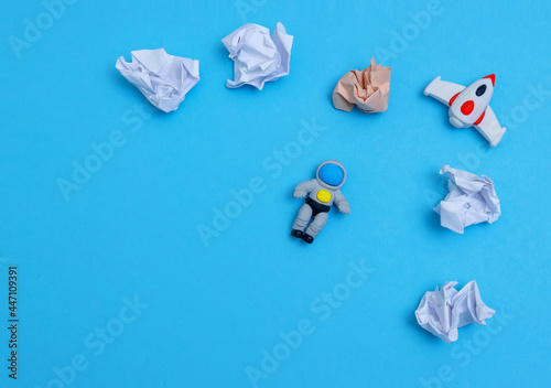 Top view of toy astronaut and crumpled paper like plates and clouds on a blue background.