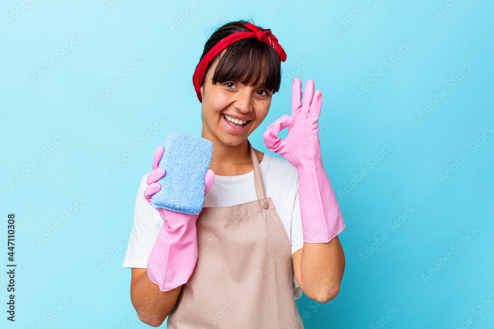 Young mixed race woman cleaning home isolated on blue background cheerful and confident showing ok gesture. Young mixed race woman cleaning home isolated on blue background cheerful and confident showing ok gesture.