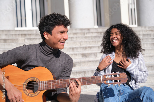 A young latin couple playing the guitar and laughing sitting on stairs. Millennial generation.