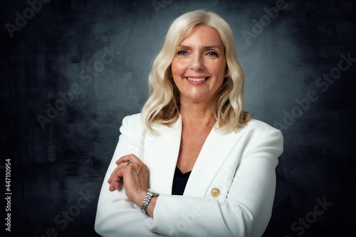 Beautiful blond haired senior womam wearing white blazer while standing at isolated dark background photo