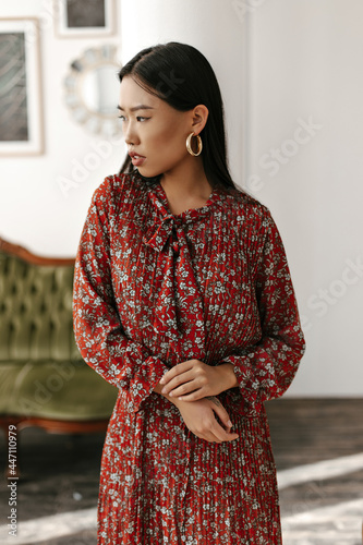 Young brunette woman in red floral dress looks into left and gently touches her arm. Asian lady poses in cozy light living room.