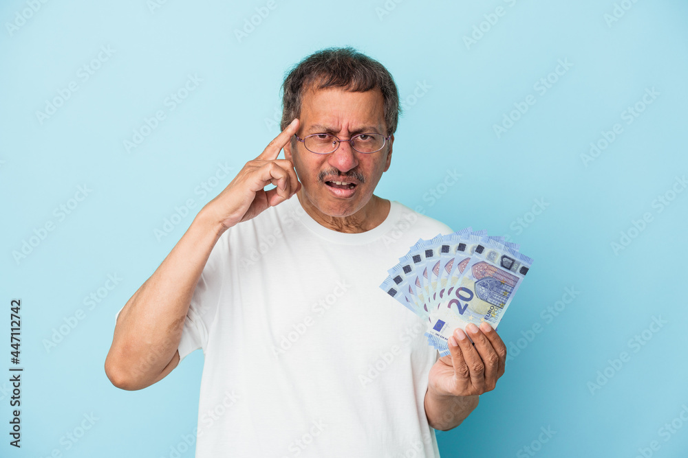 Senior indian man holding bill isolated on blue background showing a disappointment gesture with forefinger. Senior indian man holding bill isolated on blue background showing a disappointment gesture with forefinger.