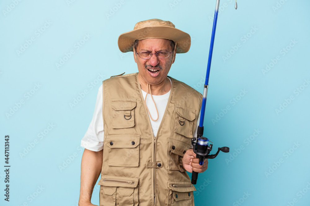 Senior indian fisherman holding rod isolated on blue background screaming very angry and aggressive. Senior indian fisherman holding rod isolated on blue background screaming very angry and aggressive.
