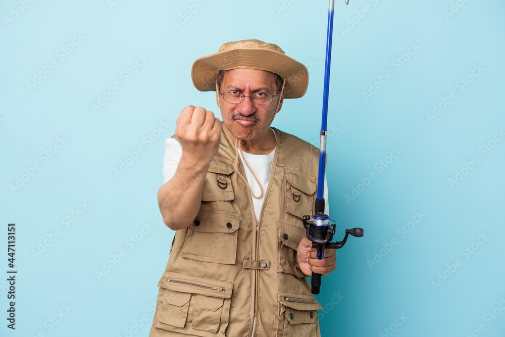 Senior indian fisherman holding rod isolated on blue background showing fist to camera, aggressive facial expression. Senior indian fisherman holding rod isolated on blue background showing fist to camera, aggressive facial expression.