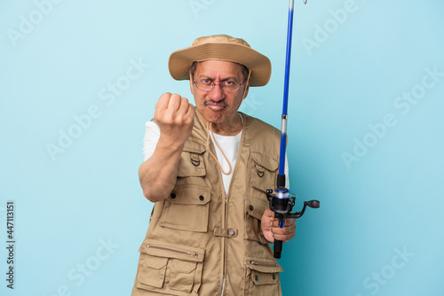 Senior indian fisherman holding rod isolated on blue background showing fist to camera, aggressive facial expression.