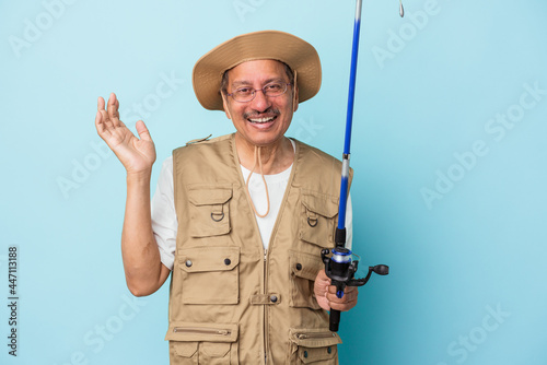 Senior indian fisherman holding rod isolated on blue background receiving a pleasant surprise, excited and raising hands.