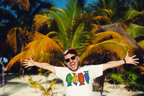 Funny handsome bearded man in white t shirt and sunglasses, gesturing triumph with raised hands against palm trees background © Davidovici