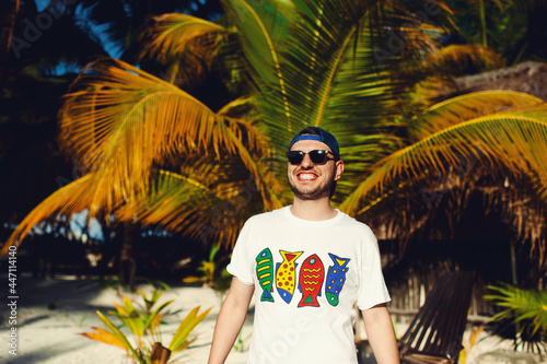 Cheerful attractive bearded tourist boy in white t shirt and sunglasses, standing while smiling broadly against palm trees background © Davidovici