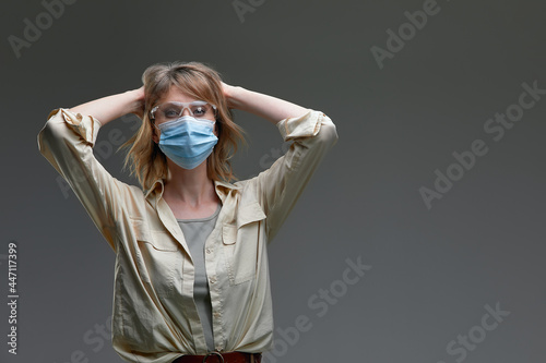 Woman in mask and glasses holding her head with her hands in panic, covid 19 concept, gray background, copy space