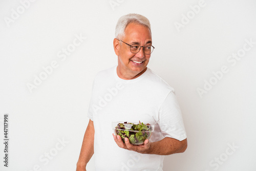 Senior american man eating salad isolated on white background looks aside smiling, cheerful and pleasant.