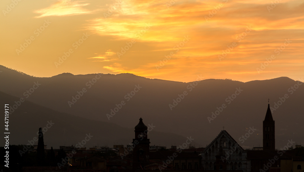 The rooftops and church towers of Pisa, Italy, silhouetted by a beautiful sunrise, with the mountains in the background