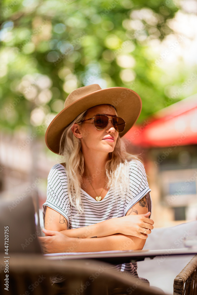 Beautiful stylish adult female in casual wear, modern hat and sunglasses sitting at table on cafe terrace.