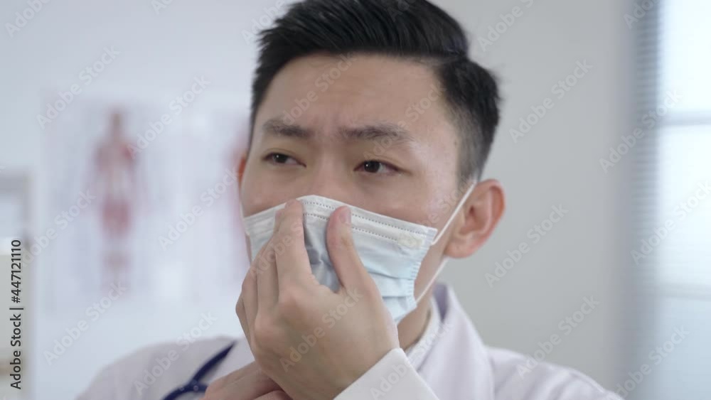 headshot asian male doctor putting on the face mask carefully and gazing into the distance with a serious look at the clinic. headshot asian male doctor putting on the face mask carefully and gazing into the distance with a serious look at the clinic.
