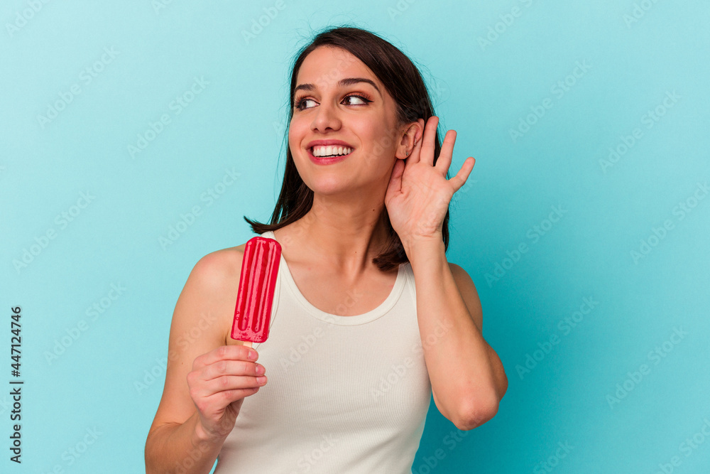Young caucasian woman holding an ice cream isolated on blue background trying to listening a gossip. Young caucasian woman holding an ice cream isolated on blue background trying to listening a gossip.