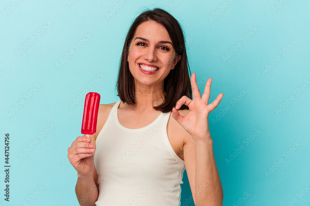 Young caucasian woman holding an ice cream isolated on blue background cheerful and confident showing ok gesture. Young caucasian woman holding an ice cream isolated on blue background cheerful and confident showing ok gesture.