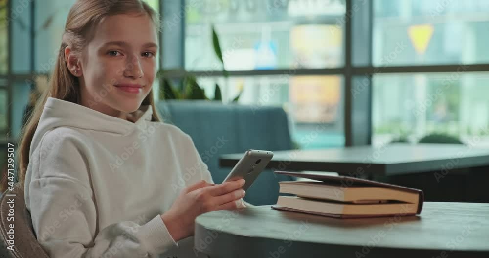 Portrait smiling preteen girl 10s using cell phone inside cafe, child looking at camera Portrait smiling preteen girl 10s using cell phone inside cafe, child looking at camera
