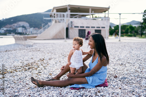 Mom with a little girl are sitting on a pebble beach by the sea