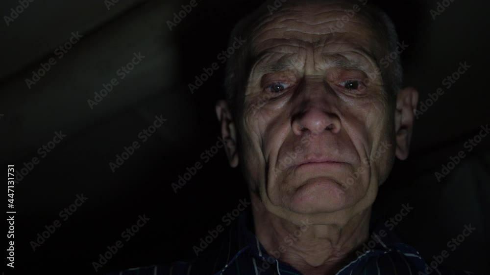 Serious aged man with wrinkles looks straight with concentrated face under bright electric light in darkness at home close view
