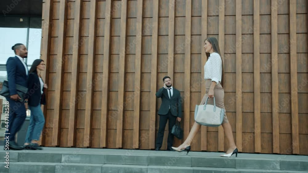 Side view of beautiful young businesswoman walking outdoors along modern office center wearing elegant clothing and meeting people in formal clothing Side view of beautiful young businesswoman walking outdoors along modern office center wearing elegant clothing and meeting people in formal clothing