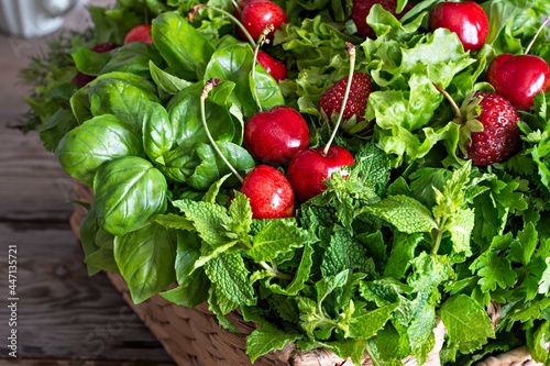 Fresh various fragrant herbs with cherries and strawberries in a wicker basket on a wooden background. Healthy food concept. Summer garden harvest.