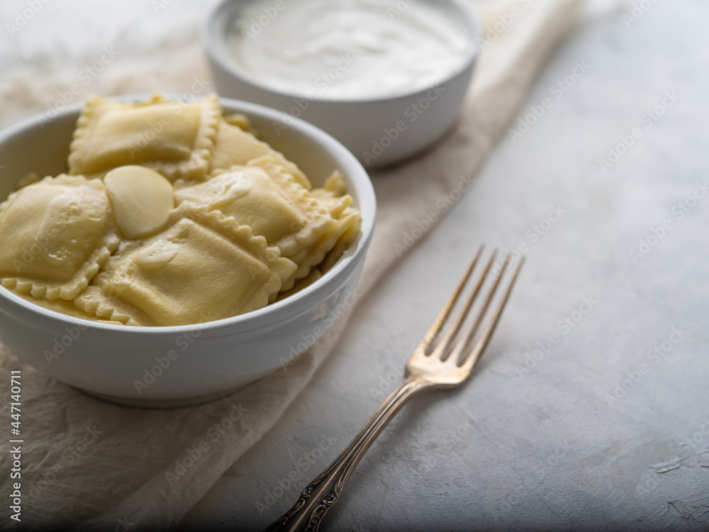 Ravioli in a white bowl and sour cream. There is a fork nearby. Delicious and healthy food. Home and restaurant cuisine. White background. High angle view. Ravioli in a white bowl and sour cream. There is a fork nearby. Delicious and healthy food. Home and restaurant cuisine. White background. High angle view.