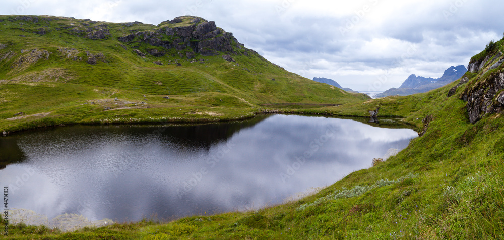 The sky is reflected in a circular mountain lake surrounded by green hills with mountains on the horizon. Deserted hills with rocks. Norway.