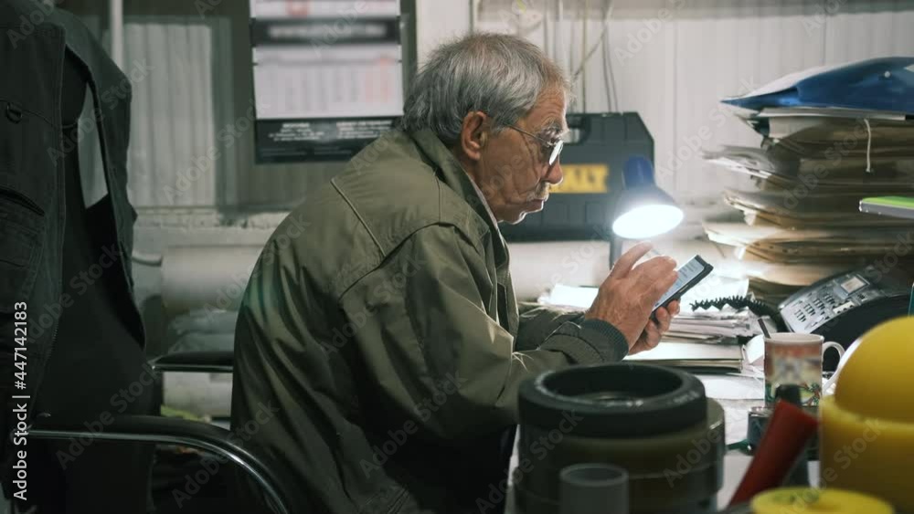 An elderly man sits at the desk looking for something on his phone. An elderly engineer swipes his phone. An aged man checks his mail and writes a message on his phone. An elderly man sits at the desk looking for something on his phone. An elderly engineer swipes his phone. An aged man checks his mail and writes a message on his phone.