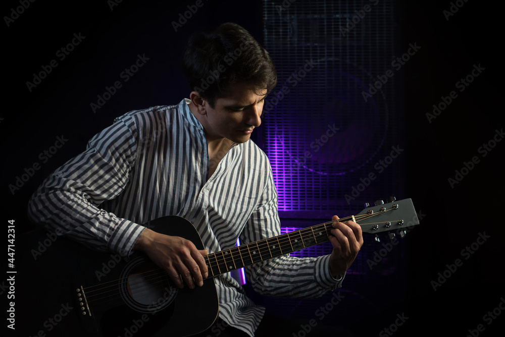 Obraz premium Photo of man sitting and playing acoustic guitar in recording studio on dark background with light effect. Repetition of the rock music of the guitarist. Rehearsal base