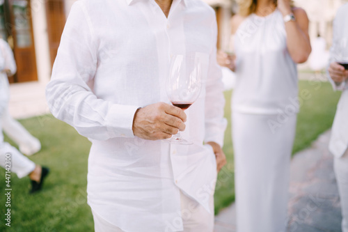 Man in a white shirt and trousers stands with a glass of red wine in his hand