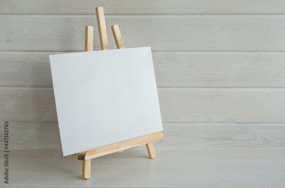 A small wooden easel with a white empty canvas on the background of a wooden wall. Creative workshop, artist's workplace, art studio.