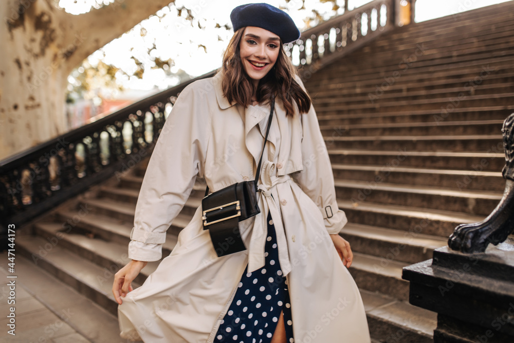 Naklejka premium Trendy young lady with brunette hair in beret, beige trench coat and crossbody bag, smiling and posing outdoors against old stairs background