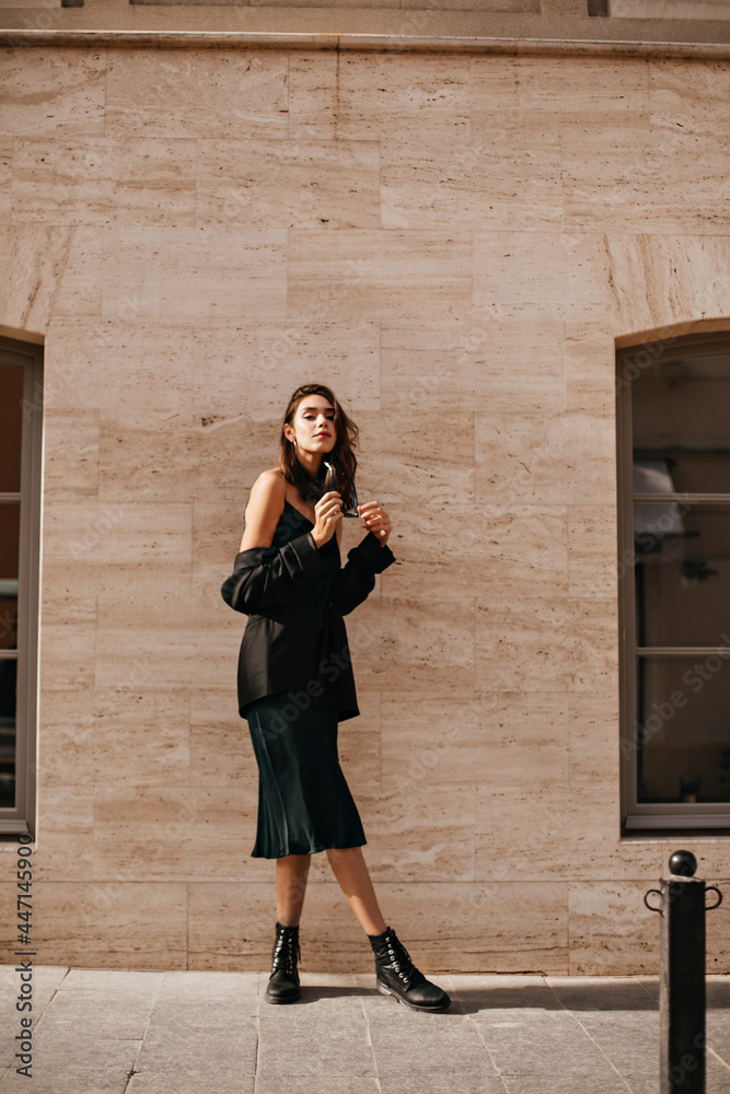 Gorgeous young brunette in midi slip dress, black oversized jacket and shoes  posing outdoors against beige wall background during sunny day Photos |  Adobe Stock
