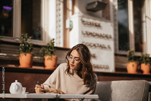 Beautiful young brunette student with makeup, glasses and beige pullover, studying, noting something and having cup of tea at sunny terrace of city cafe