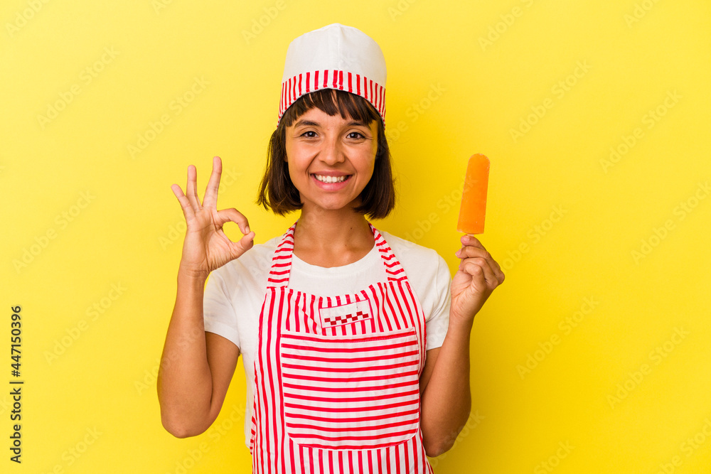 Young mixed race ice cream maker woman holding an ice cream isolated on yellow background cheerful and confident showing ok gesture. Young mixed race ice cream maker woman holding an ice cream isolated on yellow background cheerful and confident showing ok gesture.