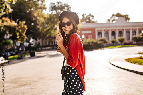 Cute young Parisienne with dark hair, beret, black sunglasses, red shirt and polka dot skirt smiling and posing in city. Old building, trees and sunny weather on background © Look!