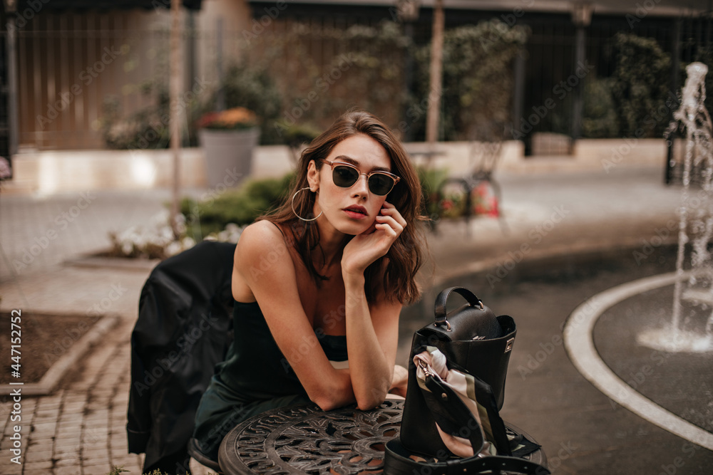 Pretty young girl with dark wavy hairstyle, red lips and black sunglasses,  wearing slip green dress posing on cafe terrace outdoors during daytime  Stock Photo | Adobe Stock